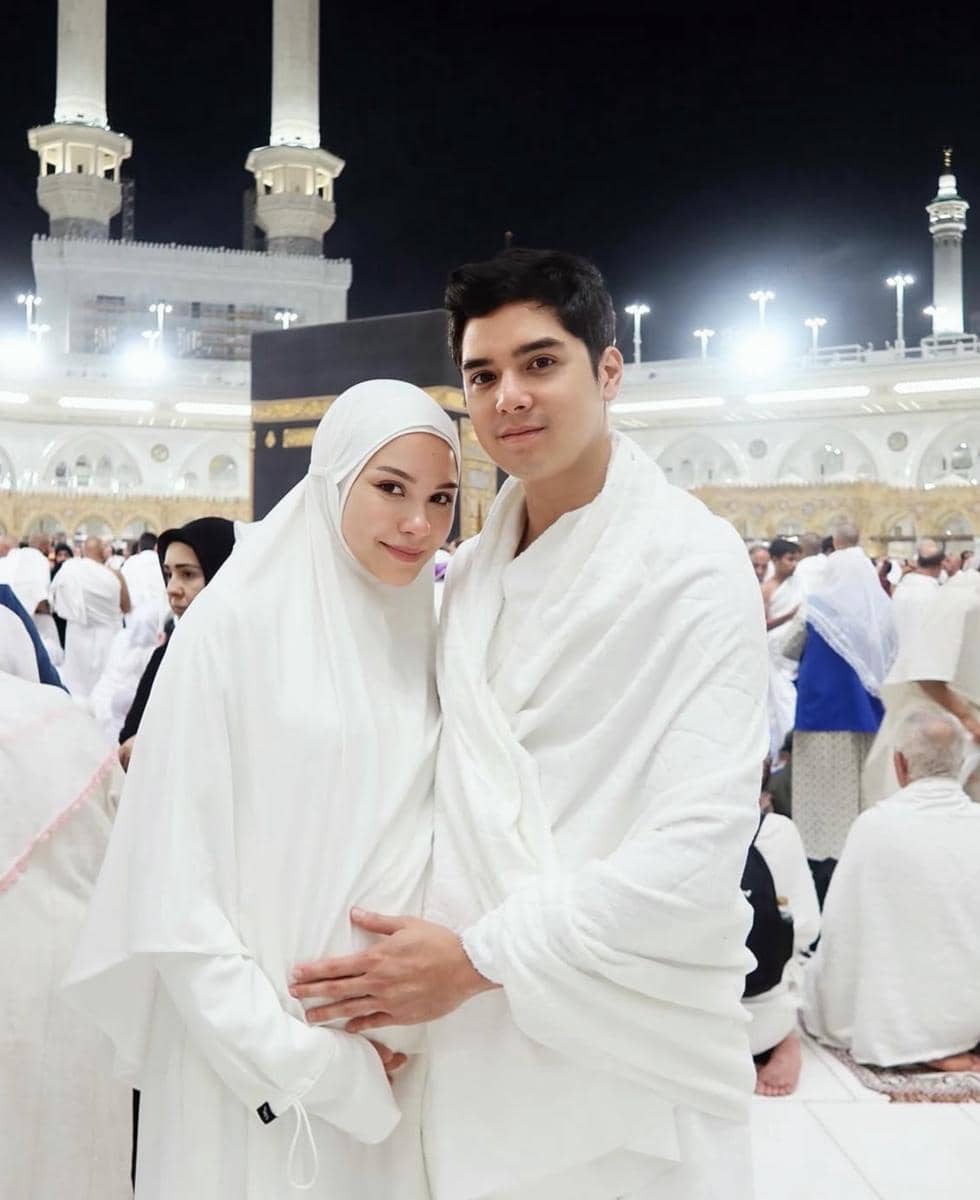 Sepasang suami istri mengenakan pakaian ihram berwarna putih berpose di depan Ka'bah di Masjidil Haram pada malam hari.