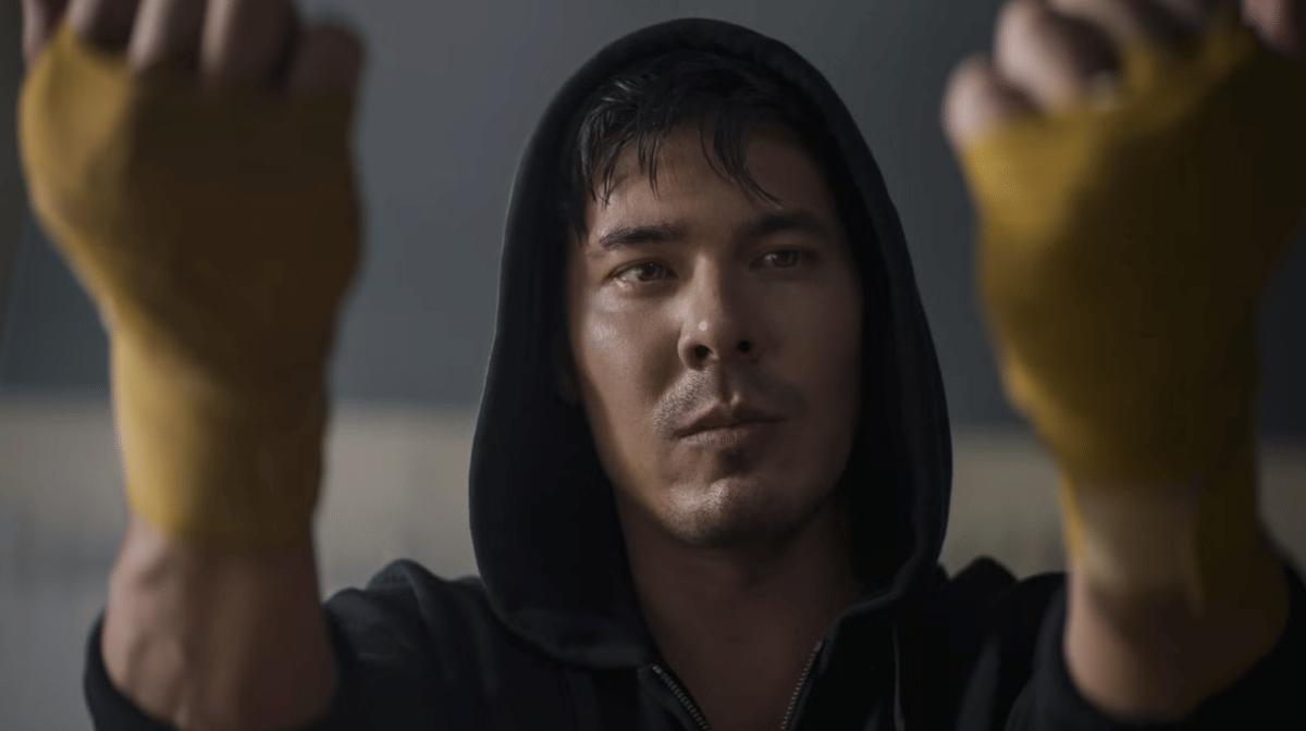 Seorang pria mengenakan hoodie hitam dengan perban kuning di tangannya tampak fokus menatap ke depan di ruangan redup.