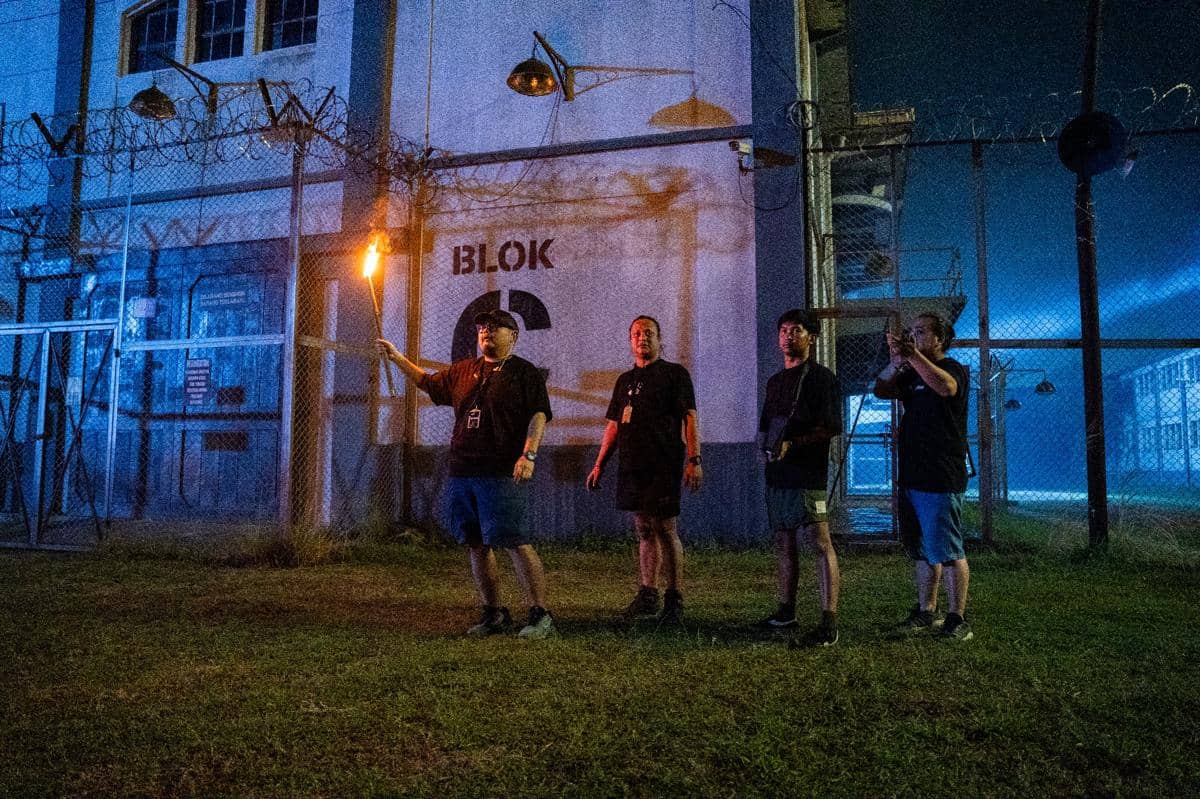 Empat kru produksi berdiri di depan bangunan bertuliskan Blok C pada malam hari, salah satu memegang obor di area berpagar kawat berduri.