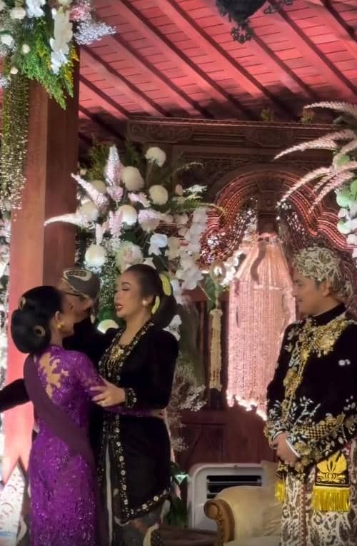 Beberapa tamu mengenakan busana adat Jawa berwarna hitam dan ungu saling berpelukan di pelaminan berhias bunga putih dan ungu.