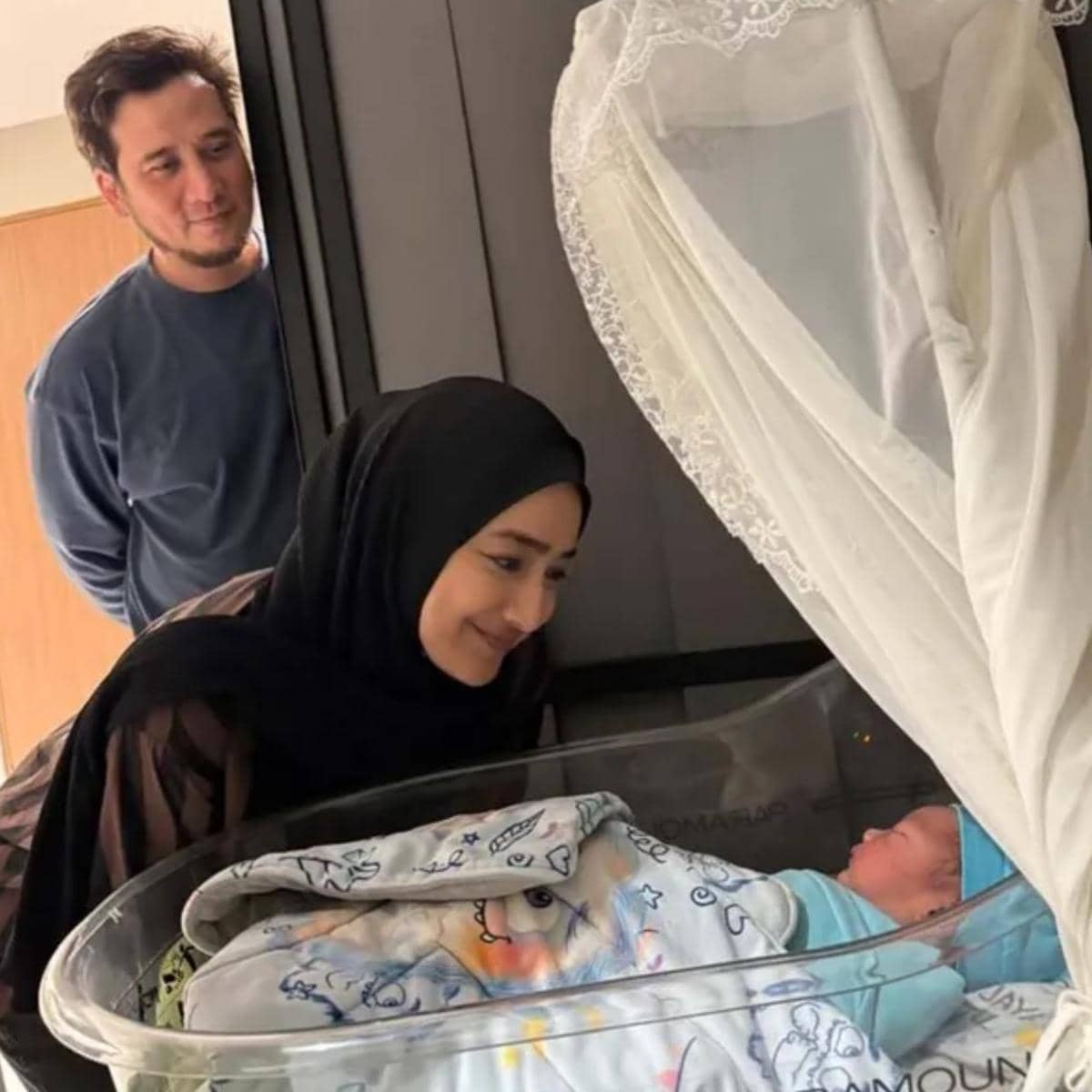 Seorang wanita berhijab tersenyum melihat bayi yang terbaring di boks bayi, sementara seorang pria berdiri di belakangnya memperhatikan dengan lembut.
