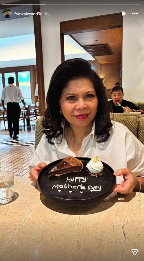 Seorang wanita tersenyum sambil memegang piring berisi kue dengan tulisan Happy Mother’s Day di sebuah restoran.