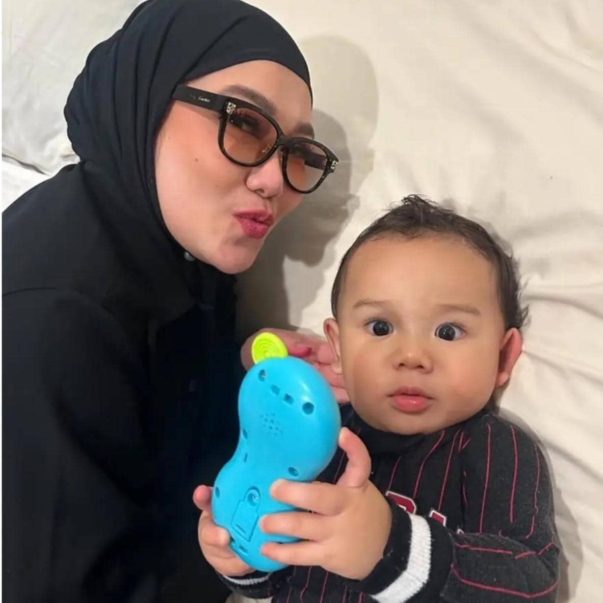 Seorang wanita berhijab mengenakan kacamata hitam berpose bersama bayi yang memegang mainan berwarna biru di atas tempat tidur.