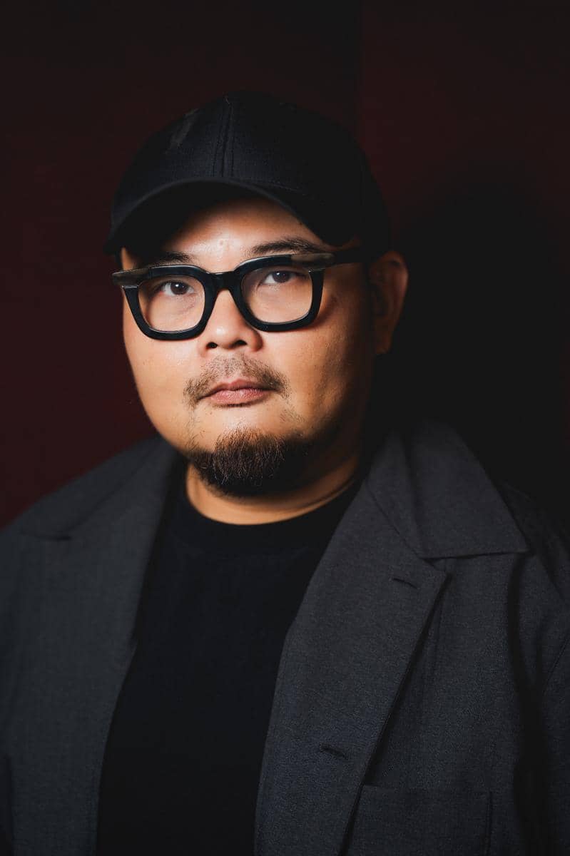 Seorang pria berkacamata hitam mengenakan topi dan jas gelap berpose di depan latar belakang berwarna gelap.