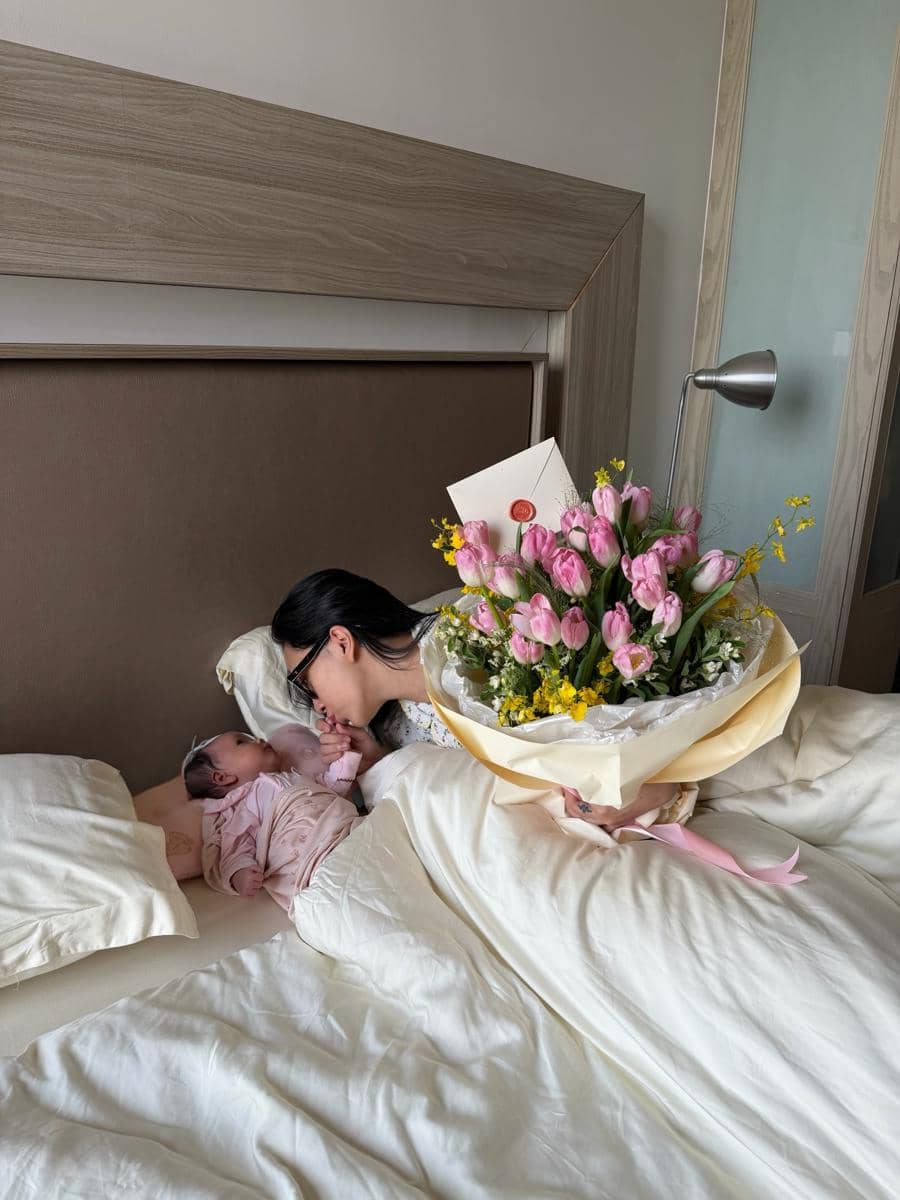 Seorang ibu berbaring di tempat tidur mencium tangan bayinya sambil memegang buket bunga berisi tulip merah muda dan bunga kuning.