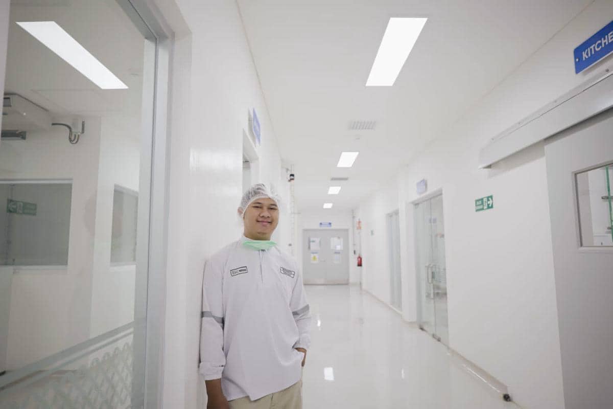 Seorang pria mengenakan pakaian laboratorium putih dan penutup kepala berdiri di koridor fasilitas bersih dengan pencahayaan terang.