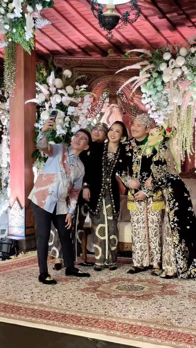 Beberapa orang berpose bersama pengantin dalam balutan busana adat Jawa di pelaminan berhias bunga putih dan ornamen kayu ukir.