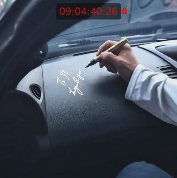 Seseorang menandatangani dashboard mobil dengan spidol emas di bagian dalam kendaraan berwarna gelap.