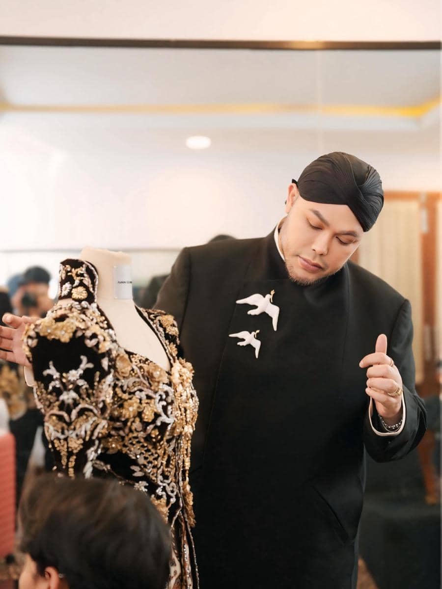 Seorang pria mengenakan pakaian hitam dengan hiasan burung putih sedang memperhatikan gaun hitam berornamen emas di atas manekin.