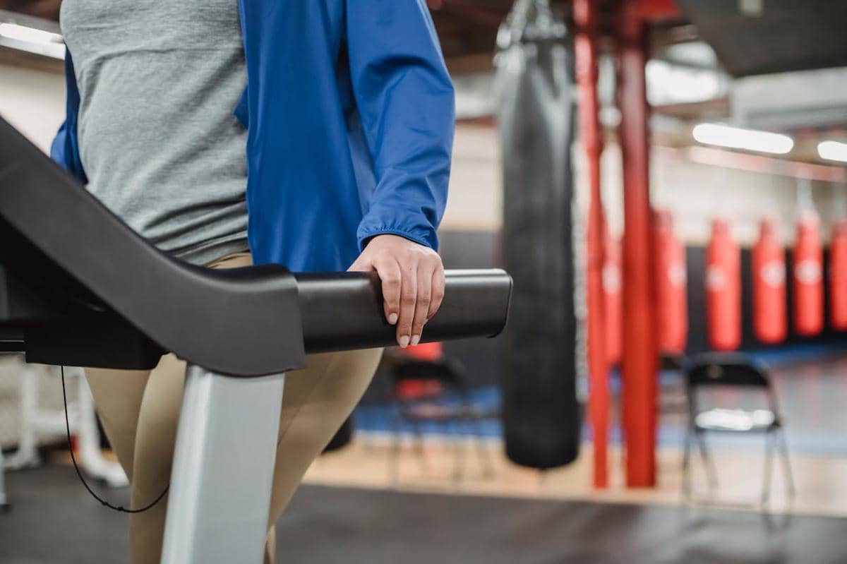 Seseorang mengenakan jaket biru dan celana krem memegang handle treadmill di gym dengan latar belakang alat tinju merah.