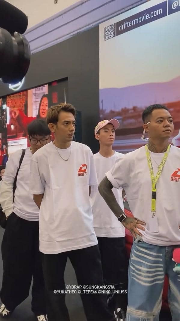 Beberapa pria mengenakan kaus putih bertuliskan logo Drift berdiri di area pameran dengan latar belakang layar besar dan spanduk promosi film.