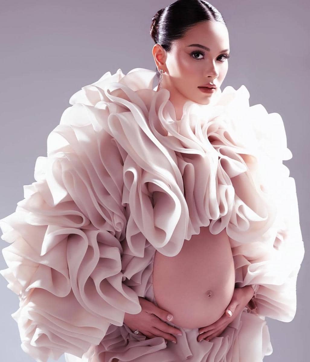 Seorang wanita hamil mengenakan gaun berlapis ruffle berwarna krem dengan gaya elegan dan memegang perutnya di latar studio lembut.
