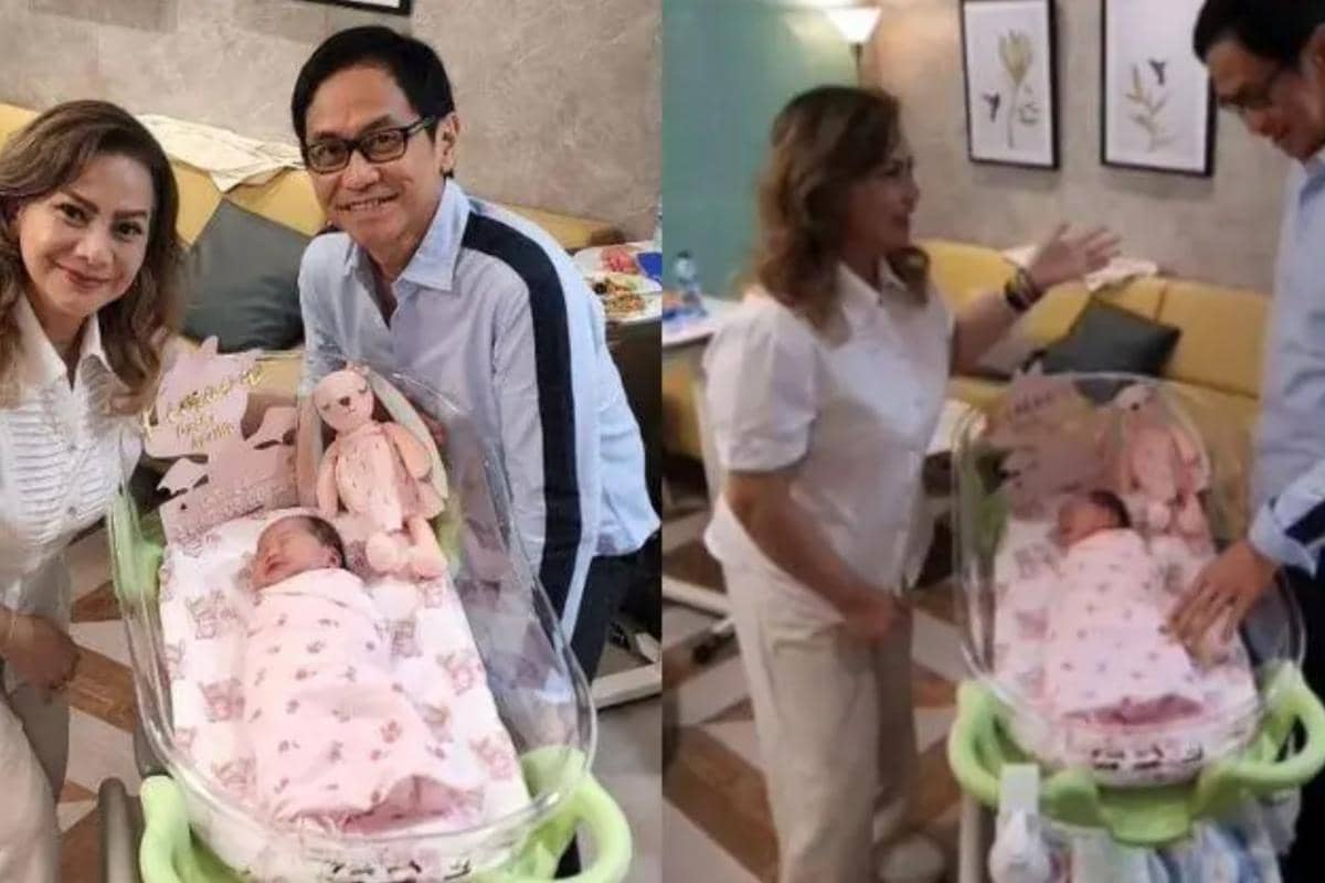 Sepasang pria dan wanita tersenyum di samping bayi yang tidur di boks bayi dengan selimut dan boneka berwarna pink di ruang tamu.