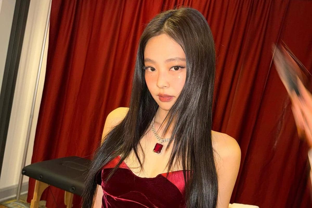 Seorang perempuan berambut panjang mengenakan gaun beludru merah dan kalung batu permata besar berwarna merah di depan tirai merah.