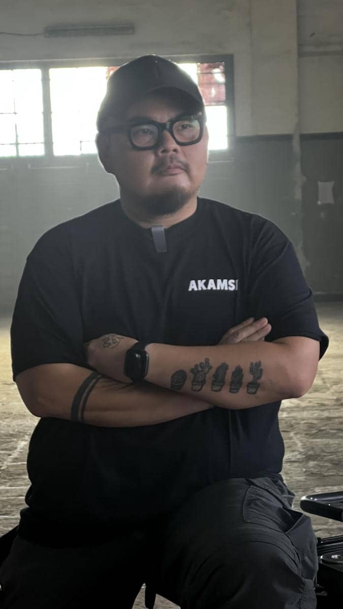 Seorang pria berkacamata dan mengenakan topi hitam berdiri dengan tangan terlipat di dalam ruangan dengan pencahayaan redup.