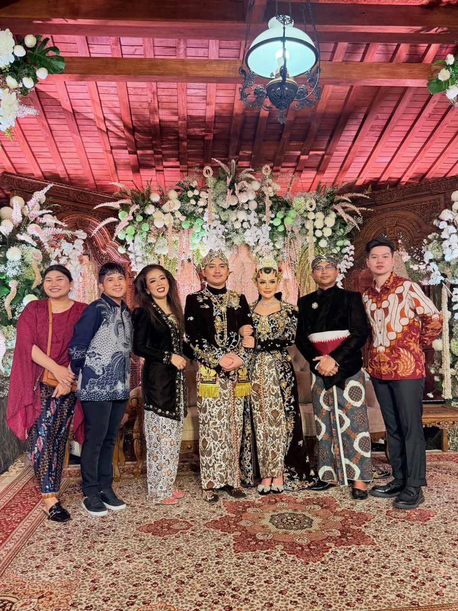 Sejumlah tamu berpose bersama pengantin dalam resepsi pernikahan bergaya tradisional Jawa dengan dekorasi bunga dan nuansa kayu.
