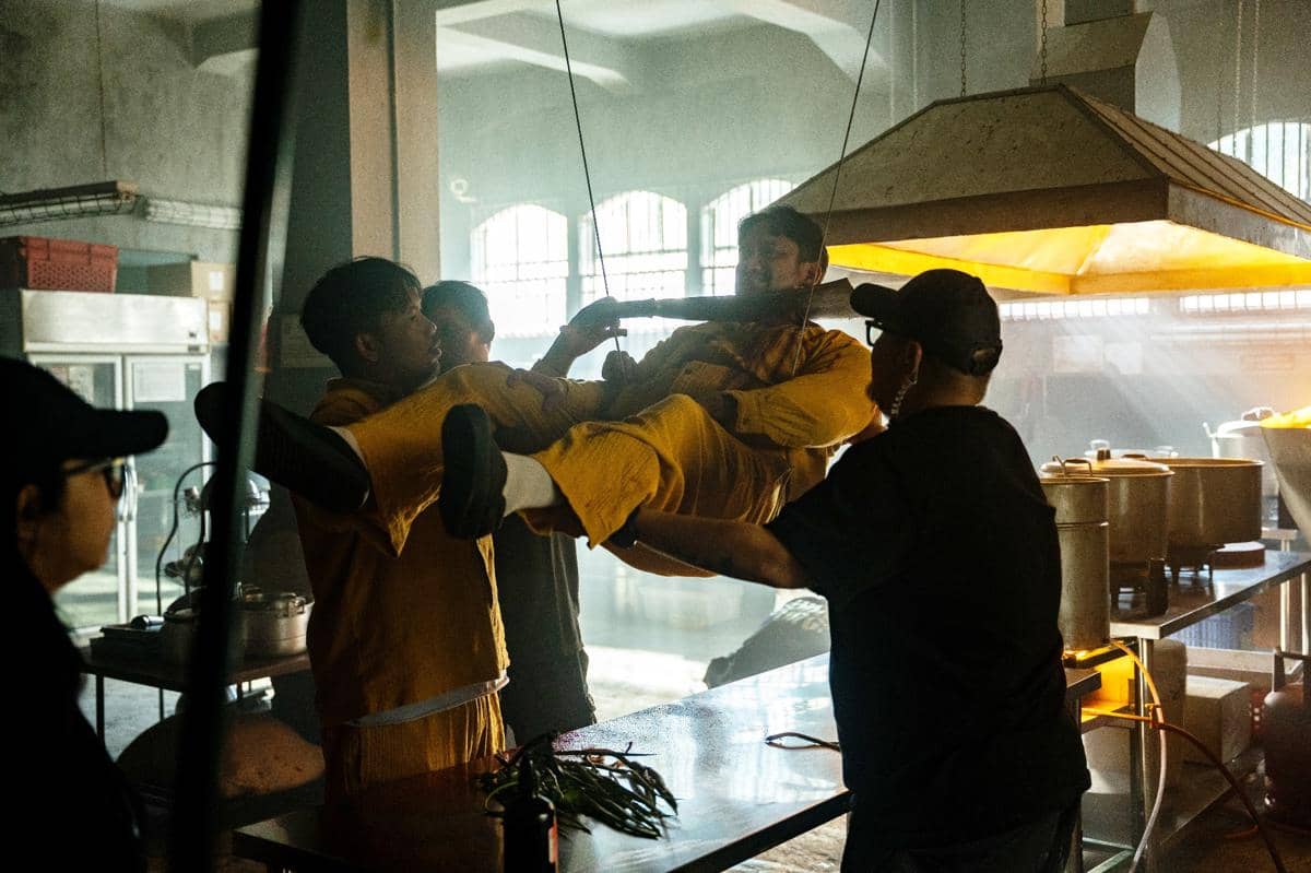 Beberapa kru film mengangkat seorang aktor dengan tali di dapur industri besar yang dipenuhi peralatan memasak dan cahaya alami.