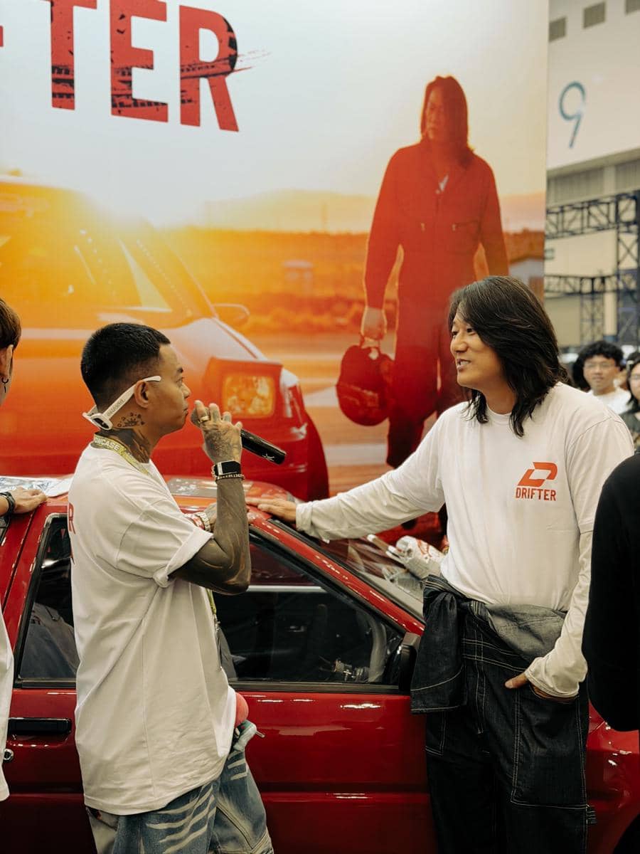 Dua pria berbincang di samping mobil berwarna merah di sebuah acara otomotif dalam ruangan dengan latar poster besar bertema balap.