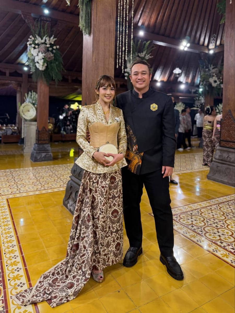 Sepasang tamu mengenakan busana tradisional Jawa berpose di resepsi pernikahan dengan latar dekorasi adat dan nuansa kayu.