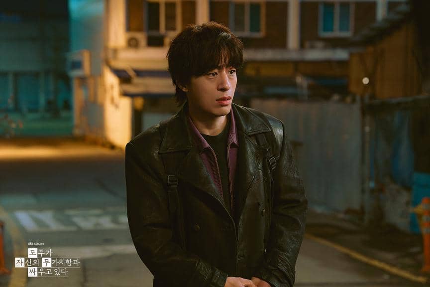 Seorang pria berjaket kulit berdiri di jalanan malam dengan ekspresi serius dalam adegan drama Korea We Are All Trying Here.