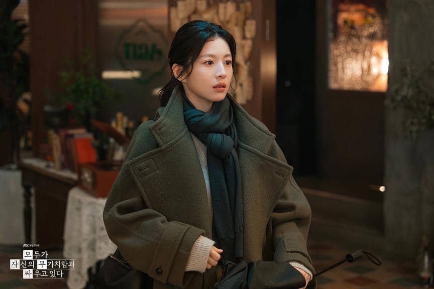 Seorang wanita mengenakan mantel tebal dan syal gelap berdiri di dalam ruangan hangat dengan pencahayaan lembut dalam adegan drama Korea We Are All Trying Here.