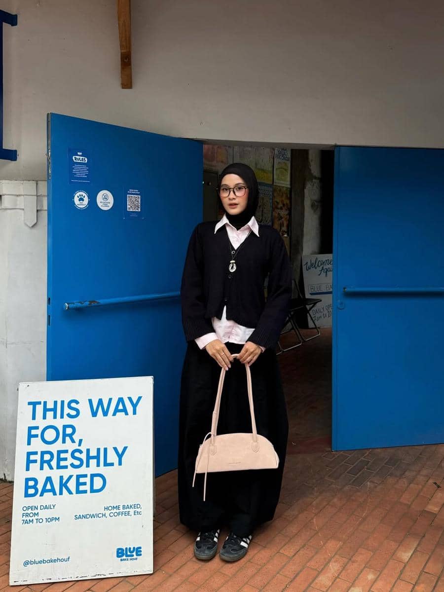 Seorang wanita berhijab mengenakan kemeja putih, sweater hitam, dan rok panjang hitam sambil memegang tas krem di depan pintu biru.