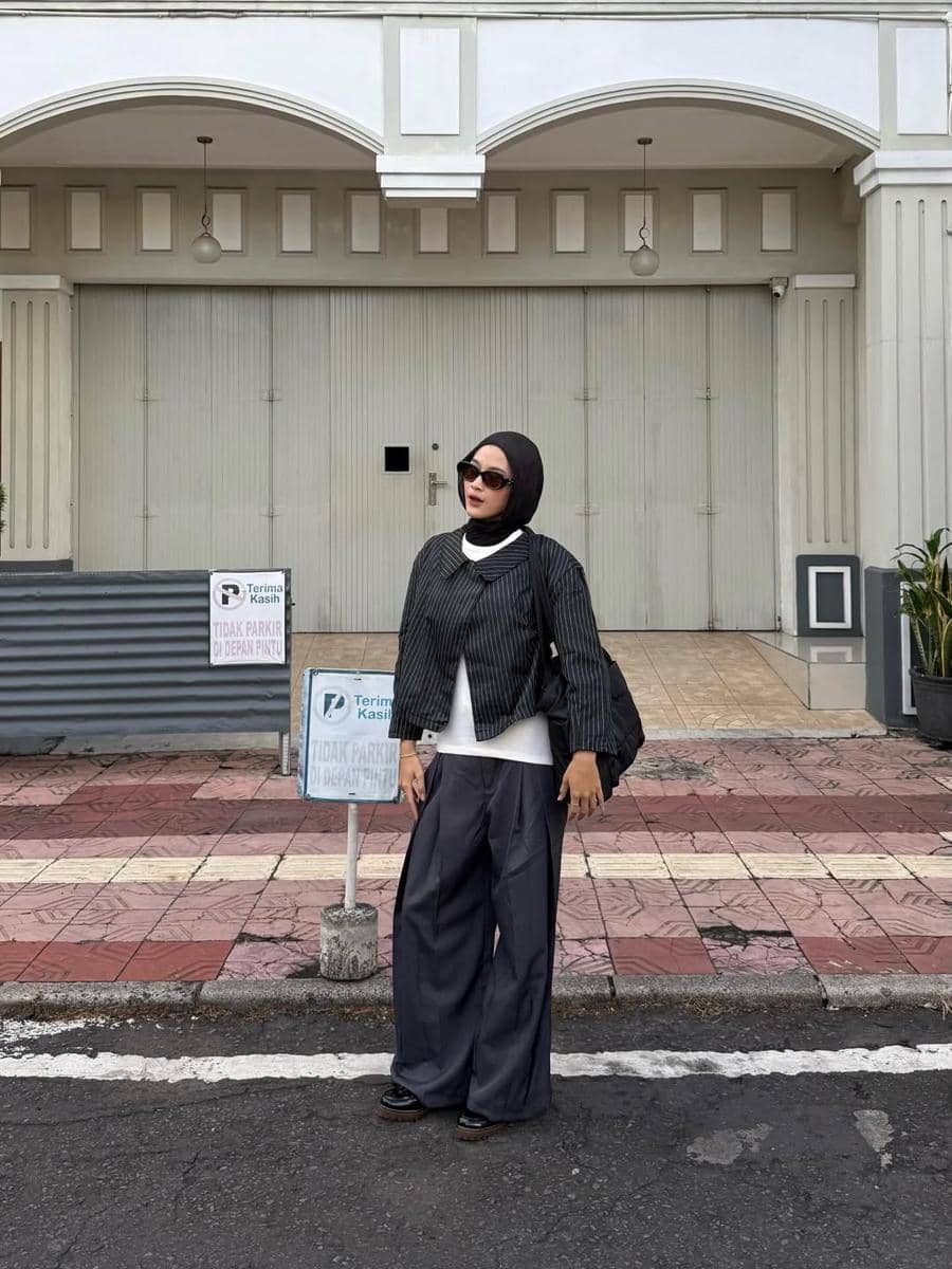 Seorang wanita berhijab mengenakan outfit bergaya kasual dengan jaket garis-garis, celana longgar abu-abu, dan kacamata hitam di depan bangunan berwarna abu.