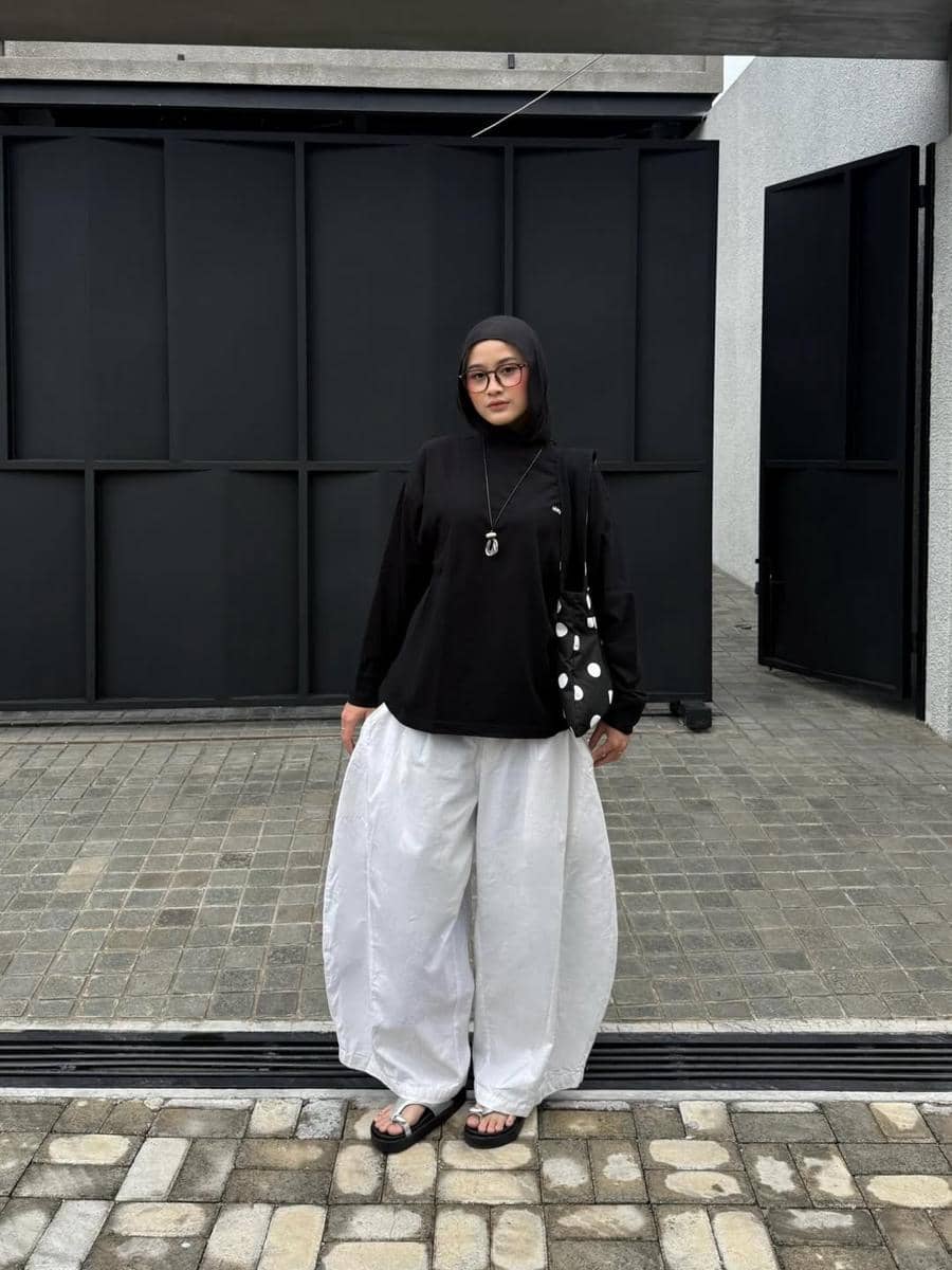 Seorang wanita berhijab mengenakan atasan hitam longgar dan celana putih lebar dengan sandal hitam, berpose di area luar bangunan modern.