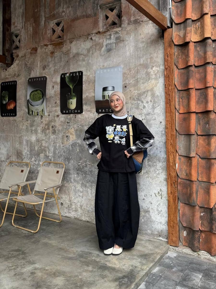 Seorang wanita berhijab mengenakan kaus hitam bergambar, kemeja kotak-kotak, dan celana kulot hitam berpose di depan dinding bergaya industrial.