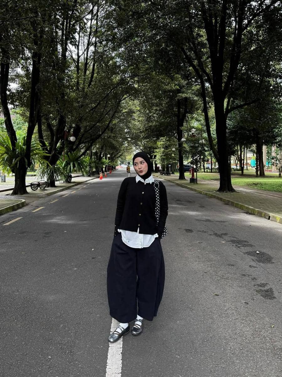 Seorang wanita berhijab bergaya kasual dengan kemeja putih, kardigan hitam, dan celana longgar hitam di jalan rindang penuh pepohonan.