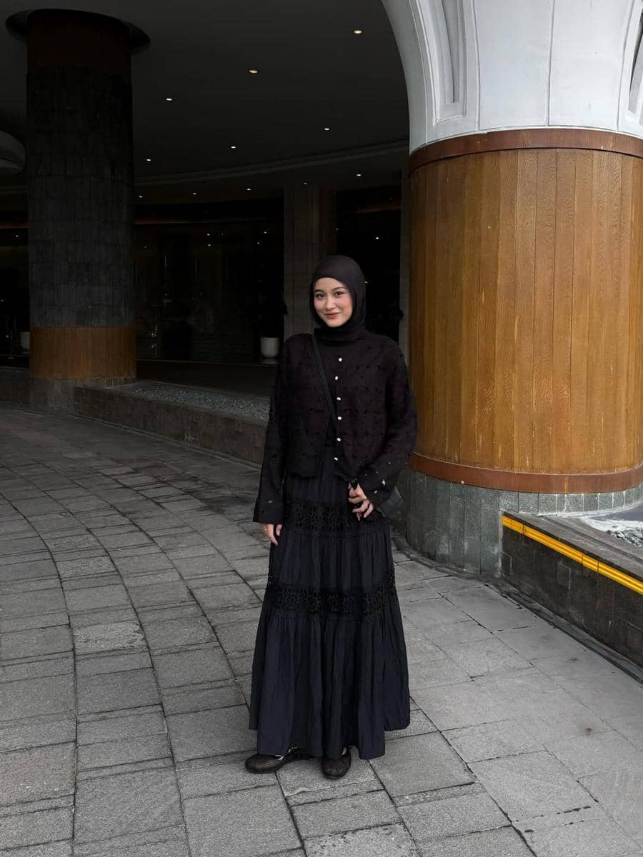 Seorang wanita berhijab mengenakan outfit serba hitam terdiri dari atasan dan rok panjang, berdiri di area luar gedung dengan pilar besar.