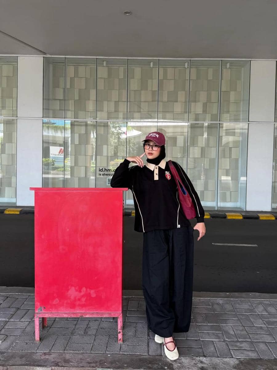Seorang wanita berhijab mengenakan topi merah marun, kemeja hitam dengan kerah krem, celana longgar hitam, dan tas bahu merah di trotoar.
