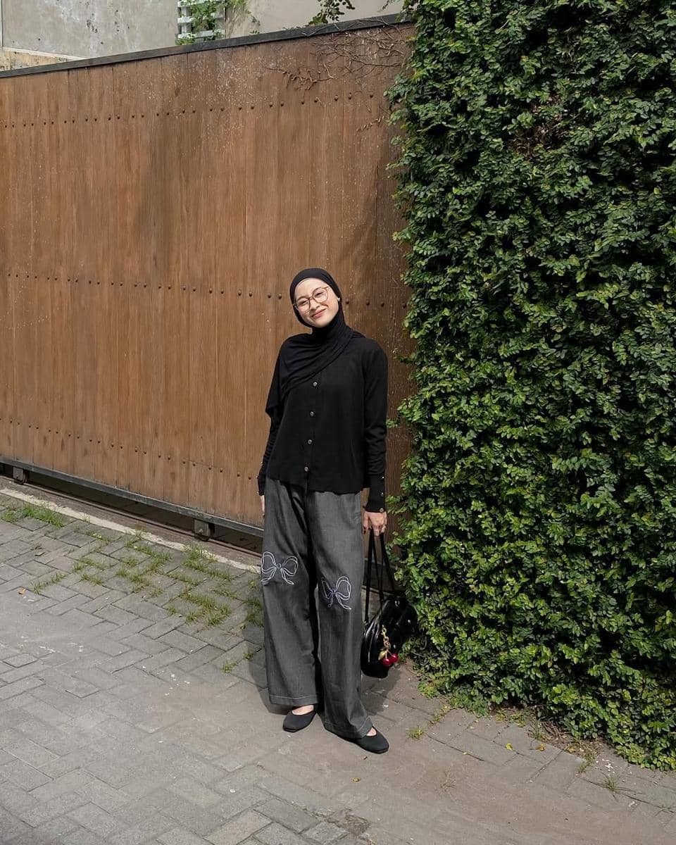 Seorang wanita berhijab mengenakan kemeja hitam dan celana abu-abu longgar dengan motif pita, berdiri di dekat dinding kayu dan tanaman merambat.