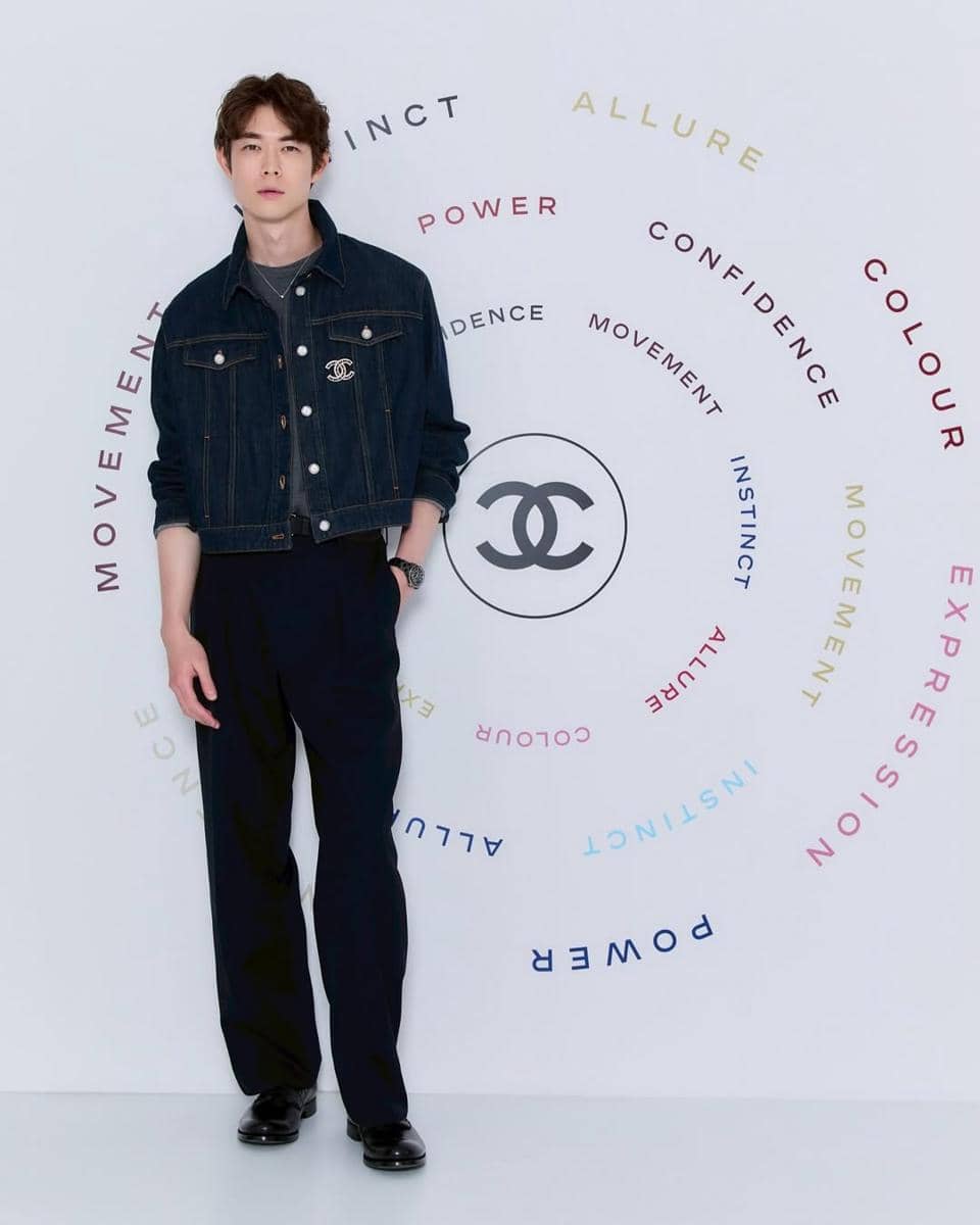 Seorang pria mengenakan jaket denim dan celana panjang hitam berdiri di depan latar bertema Chanel dengan kata-kata berwarna melingkar.