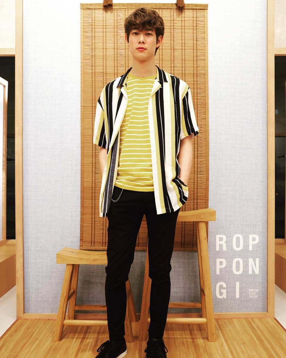 Seorang pria berdiri di dalam ruangan bergaya minimalis mengenakan kaus kuning bergaris dan kemeja motif garis vertikal hitam putih kuning.
