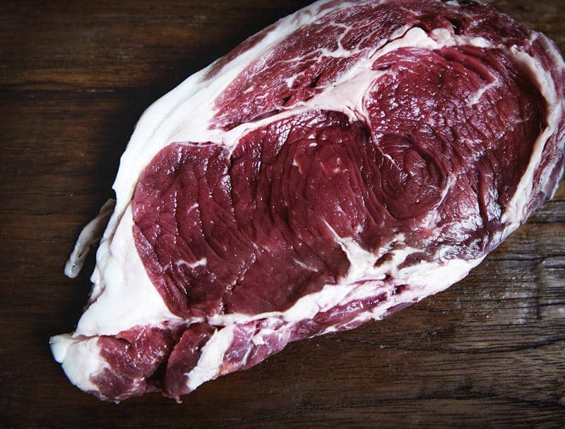 Potongan daging sapi mentah dengan lapisan lemak putih di atas permukaan meja kayu berwarna gelap.