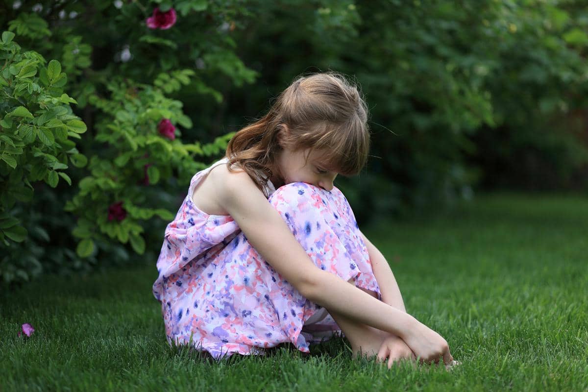 Seorang anak perempuan duduk menyendiri di taman hijau, mengenakan gaun bermotif bunga berwarna lembut dengan latar pepohonan rimbun.