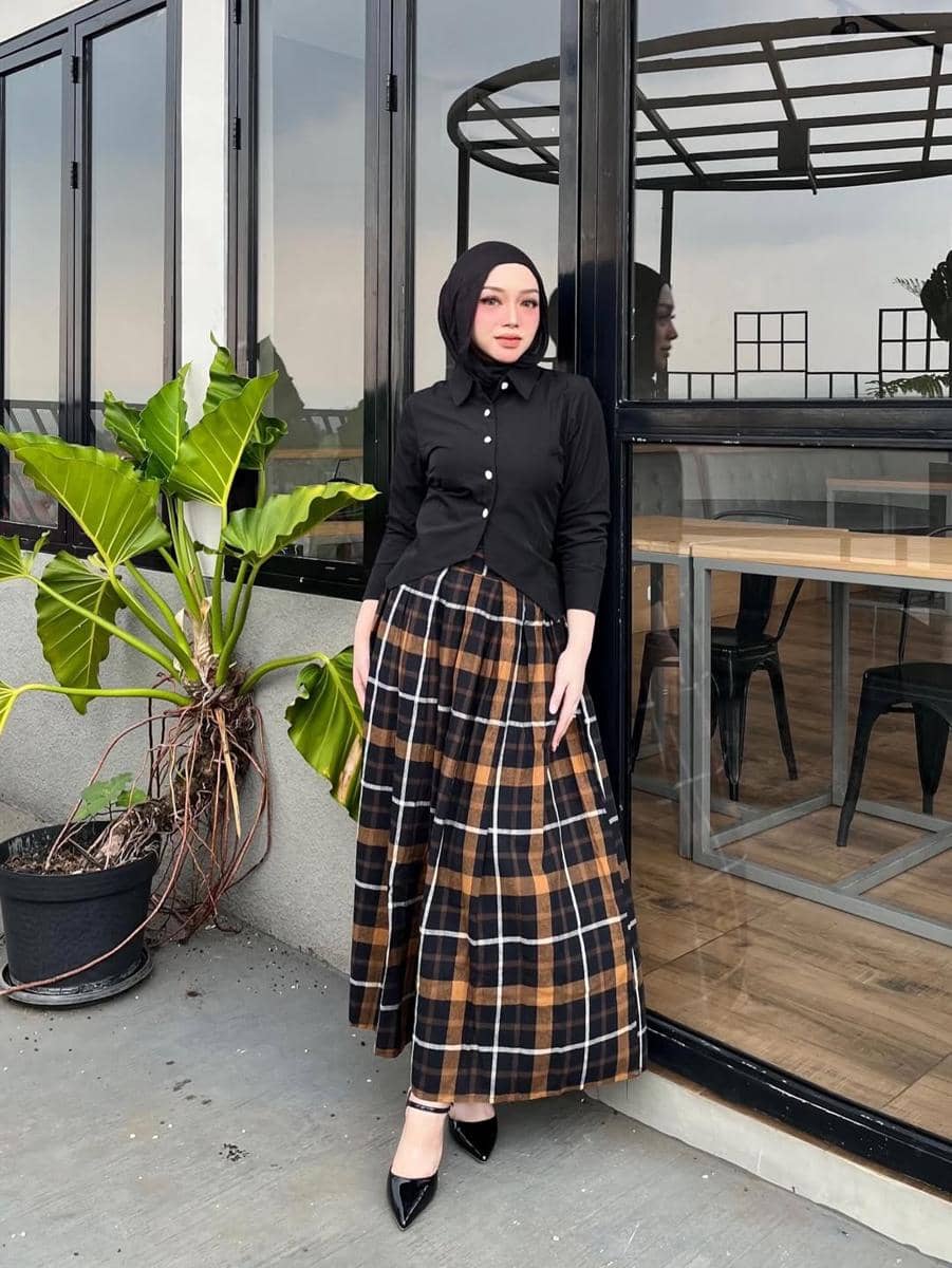 Perempuan berhijab mengenakan atasan hitam dan rok panjang motif kotak-kotak cokelat berdiri di depan kafe dengan dinding kaca.