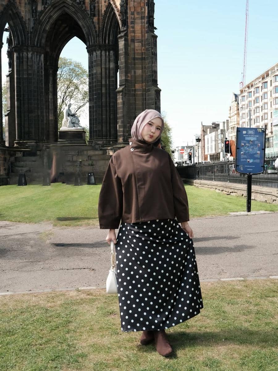 Seorang wanita berhijab mengenakan atasan cokelat dan rok hitam bermotif polkadot berpose di depan bangunan bergaya gotik di luar negeri.