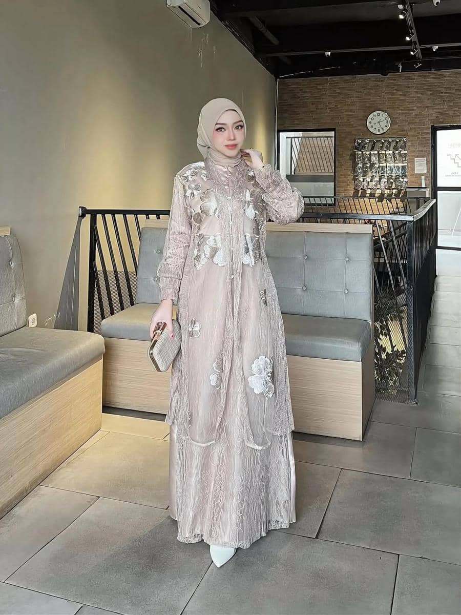 Seorang wanita berhijab mengenakan gaun kondangan berwarna pastel dengan detail renda dan bunga, memegang clutch di ruang bergaya modern.