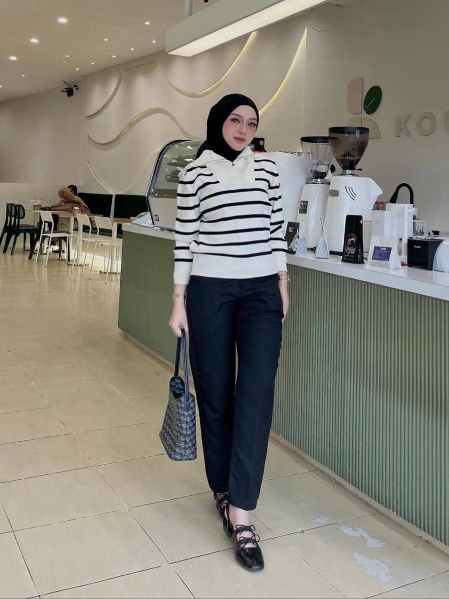 Perempuan berhijab mengenakan sweater garis hitam putih dan celana panjang hitam berpose di dalam kafe modern dengan pencahayaan hangat.