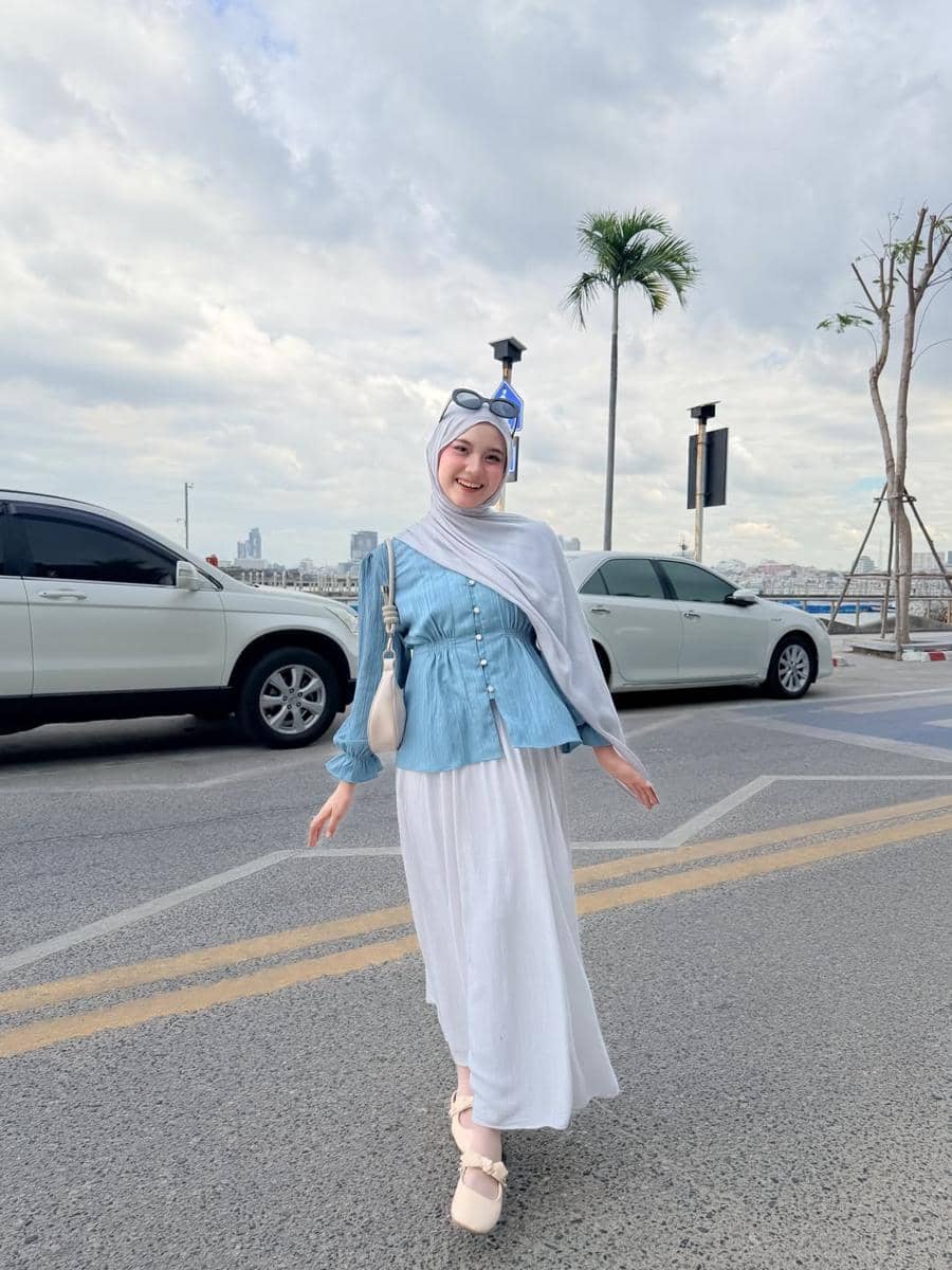 Seorang wanita berhijab bergaya kasual dengan atasan biru dan rok putih berpose di jalan dekat area parkir dengan latar langit berawan.