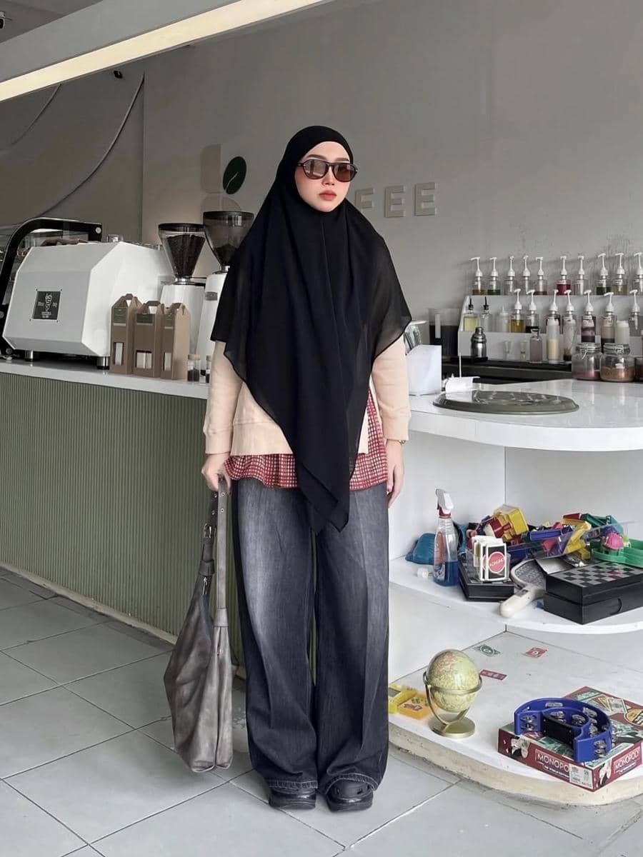 Seorang wanita berhijab hitam mengenakan kemeja kotak merah dan celana jeans longgar berdiri di kafe modern dengan dekorasi minimalis.