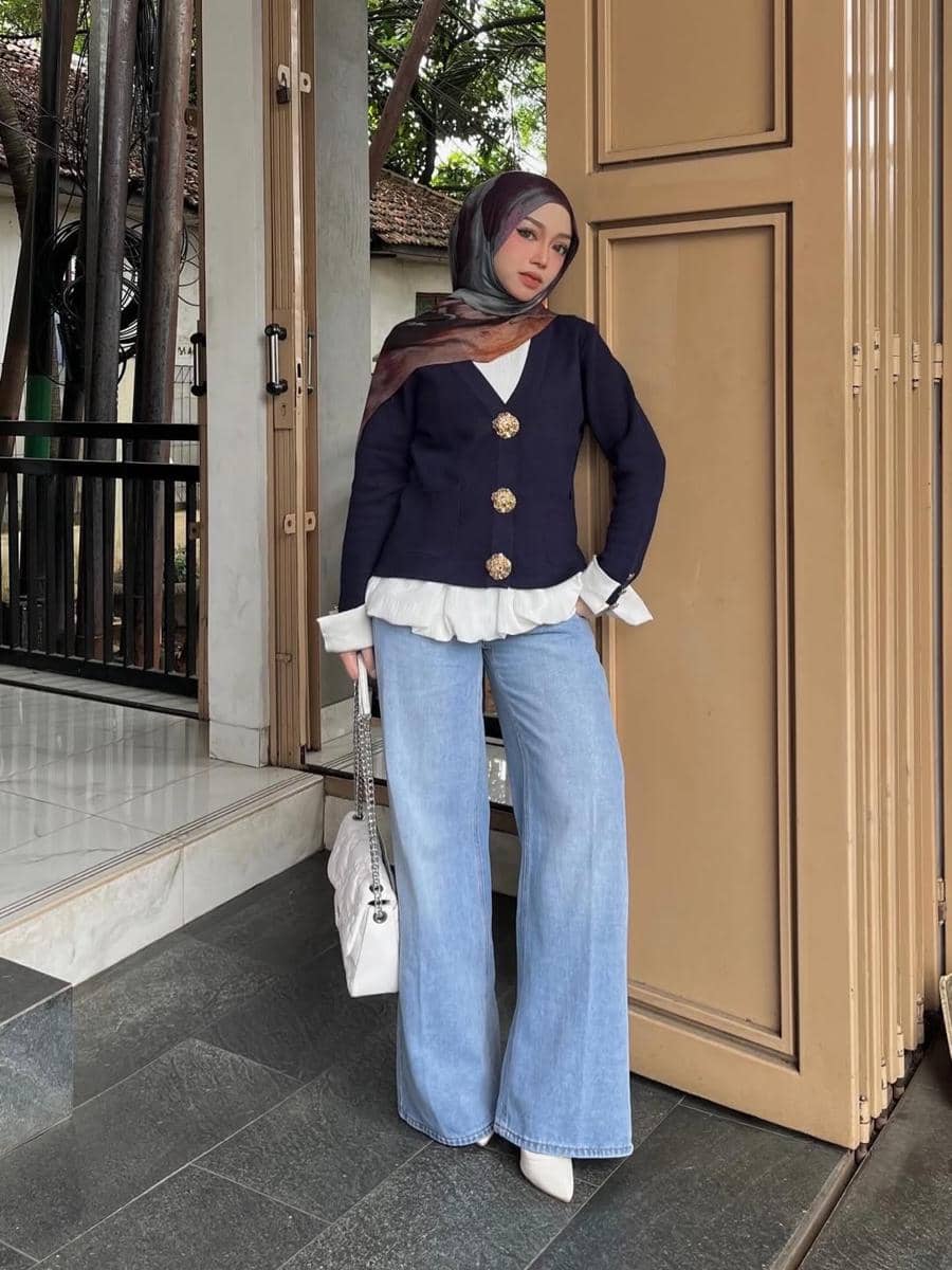 Seorang wanita berhijab mengenakan blazer biru tua, celana jeans longgar, dan tas putih berpose di depan pintu kafe bergaya modern.