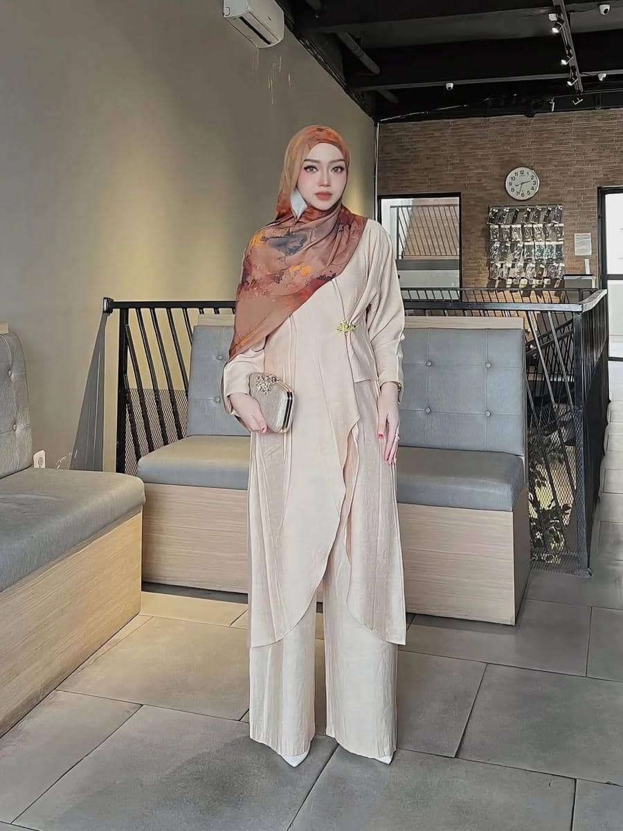 Seorang wanita berhijab mengenakan outfit kondangan berwarna krem dengan pashmina motif cokelat dan membawa clutch di ruangan bergaya modern.