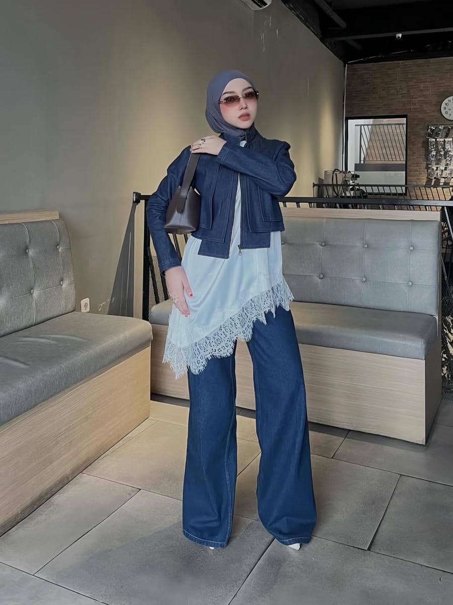 Seorang wanita berhijab bergaya kasual mengenakan jaket denim pendek, celana jeans lebar, dan kacamata hitam di dalam kafe modern.