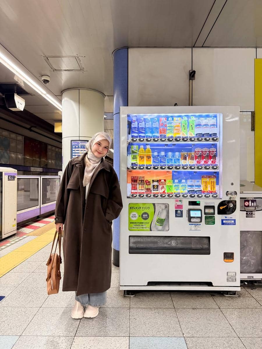 Seorang wanita berhijab mengenakan mantel cokelat panjang dan membawa tas tangan berdiri di samping mesin penjual minuman otomatis di stasiun.