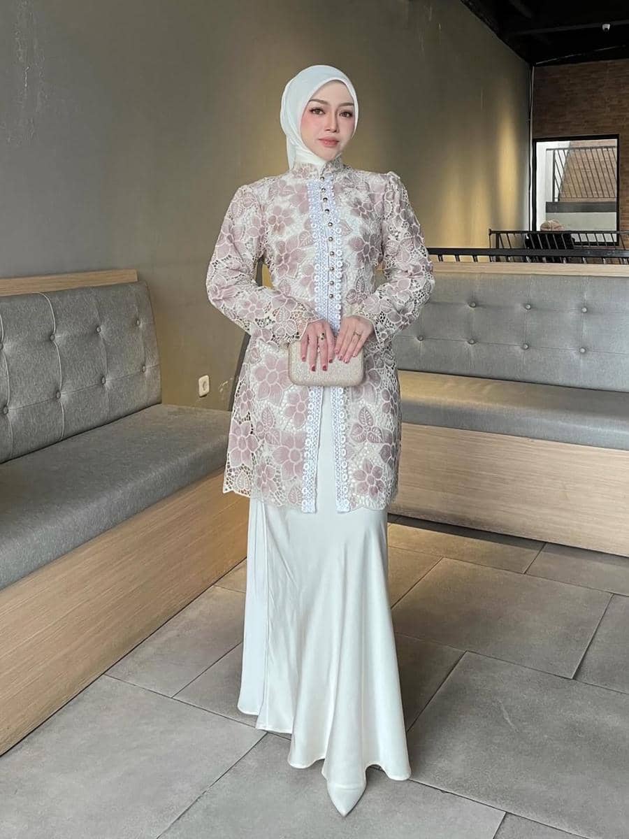 Seorang wanita berhijab mengenakan kebaya modern berwarna pastel dengan rok panjang putih dan memegang clutch di ruangan minimalis.