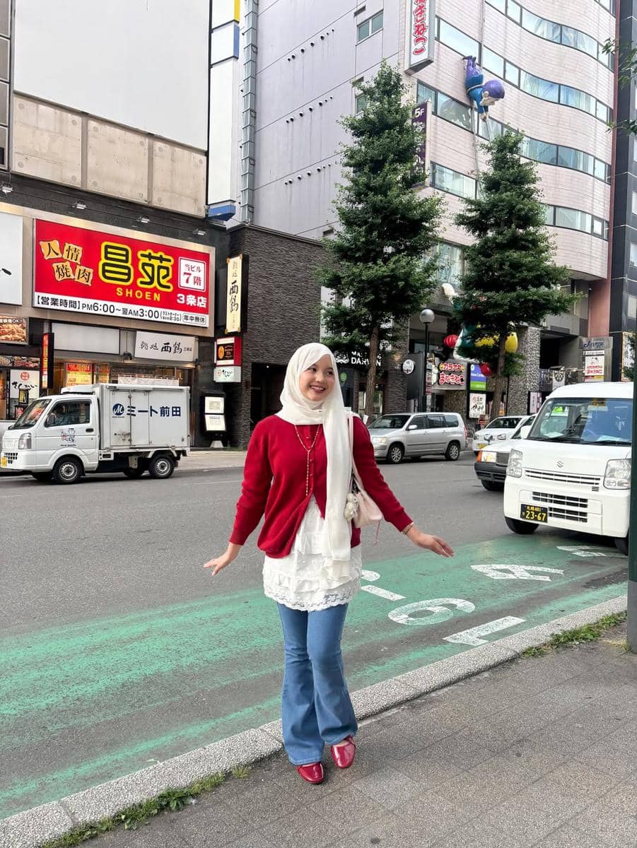 Seorang wanita berhijab mengenakan cardigan merah, tunik putih, dan celana jeans berdiri di trotoar kota dengan latar gedung dan kendaraan.