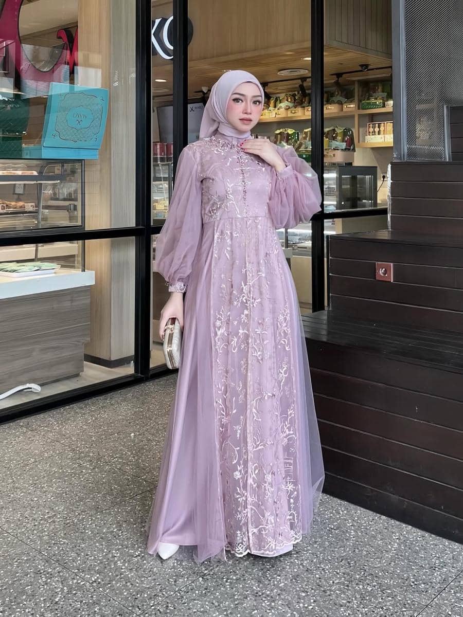 Seorang wanita berhijab mengenakan gaun panjang berwarna ungu muda dengan detail bordir bunga, berpose di depan toko kue modern.